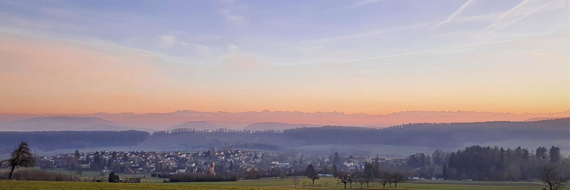 Blick auf Dettighofen und die Alpen bei Sonnenuntergang, die warmen Farben des Himmels reflektieren die ruhige Landschaft.