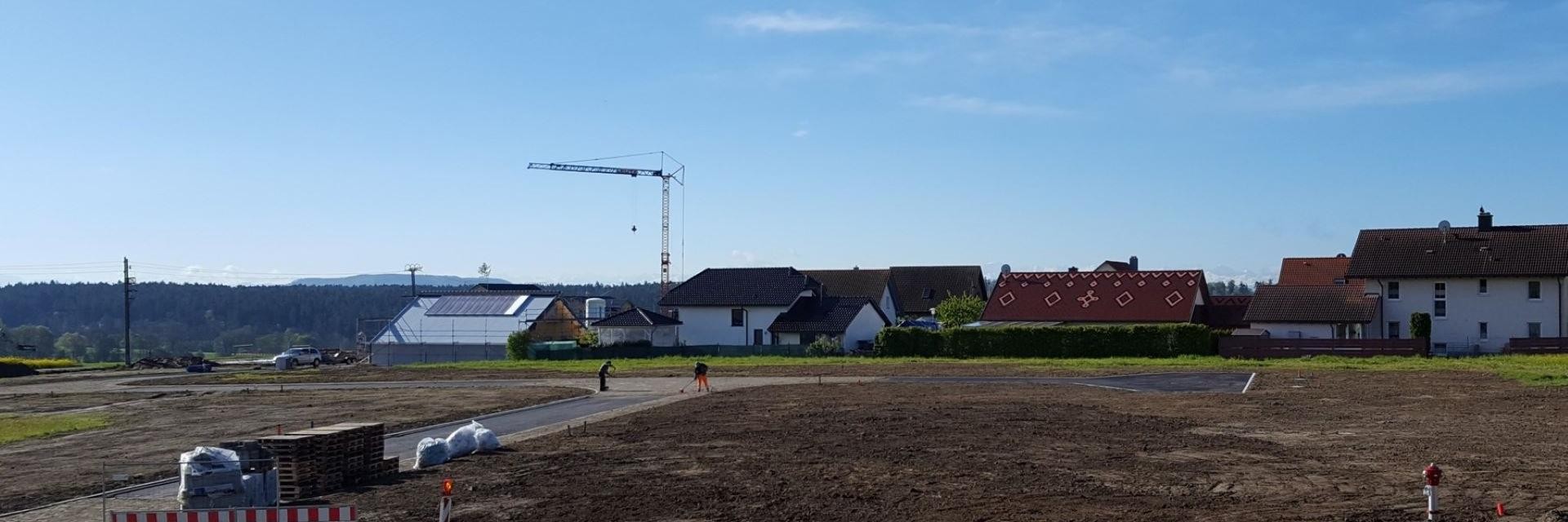 Blick auf eine Wohngegend mit mehreren Häusern und einem Baukran im Hintergrund. Im Vordergrund ist eine unbefestigte Fläche zu sehen, auf der sich einige Baustellenmaterialien befinden. Der Himmel ist klar und blau, während im Hintergrund eine Hügelkette sichtbar ist.