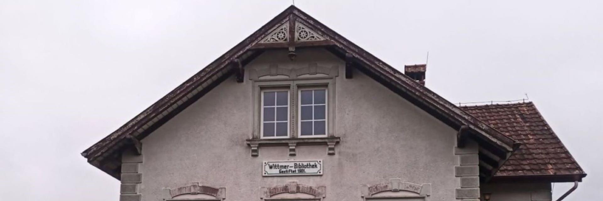 Das Bild zeigt die Fassade eines historischen Gebäudes mit einem schrägen Dach und zwei Fenstern im oberen Bereich. An der Wand befindet sich ein Schild mit der Aufschrift "Wittmer-Bibliothek". Die Fassade ist in einem hellen Farbton gehalten, während das Dach mit roten Ziegeln gedeckt ist. Der Himmel ist bewölkt, was eine gedämpfte Lichtstimmung erzeugt.