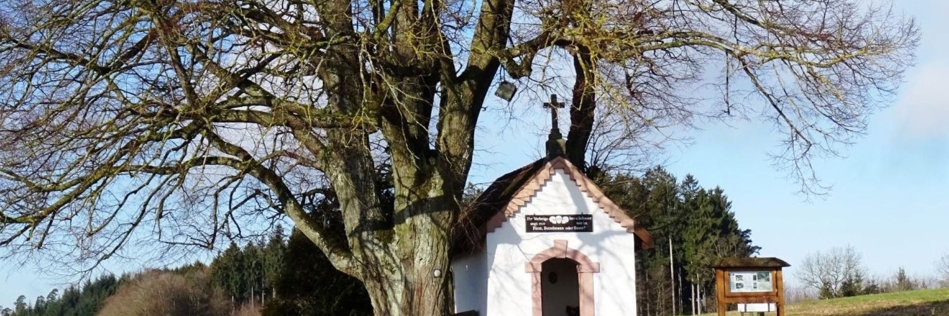 Eine kleine weiße Kapelle steht malerisch neben einem Baum, umgeben von einer idyllischen Landschaft.