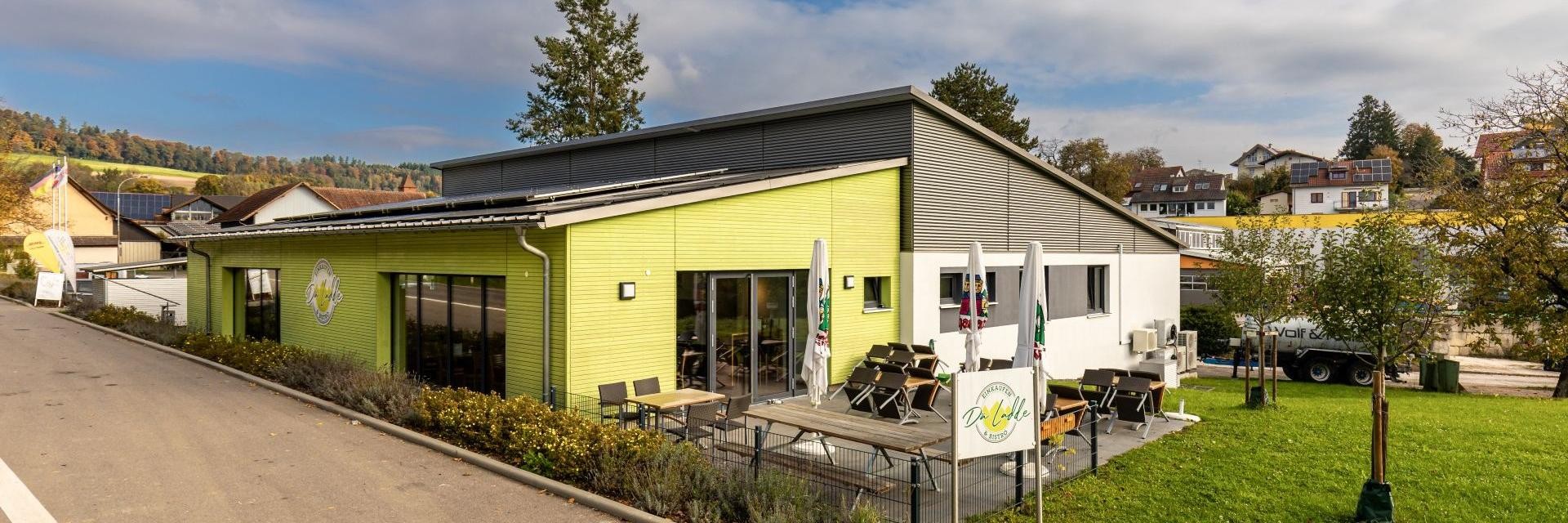 Der Dorfladen, in einem lebhaften Grünton gehalten, mit dem Außensitzbereich des Bistros im Vordergrund